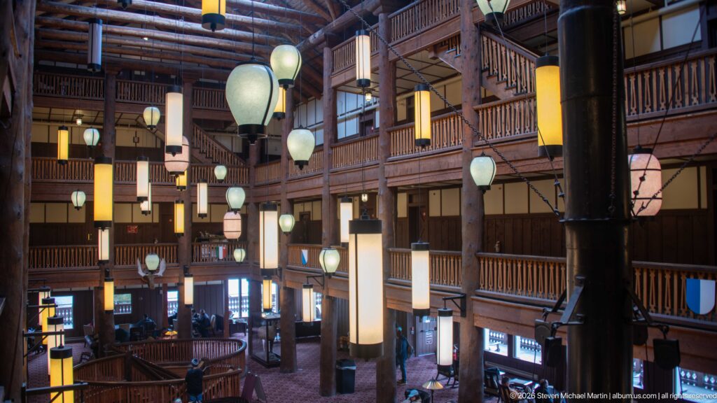 Diagonal upper-level view of the Great Room with stacked balconies, log columns, hanging lanterns, and lobby below.