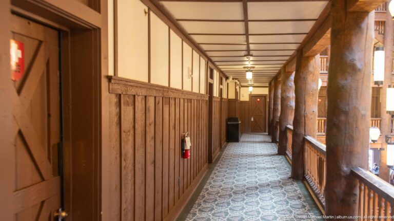 Long carpeted hotel corridor with guest-room doors, wood paneling, and log columns along the open balcony side.