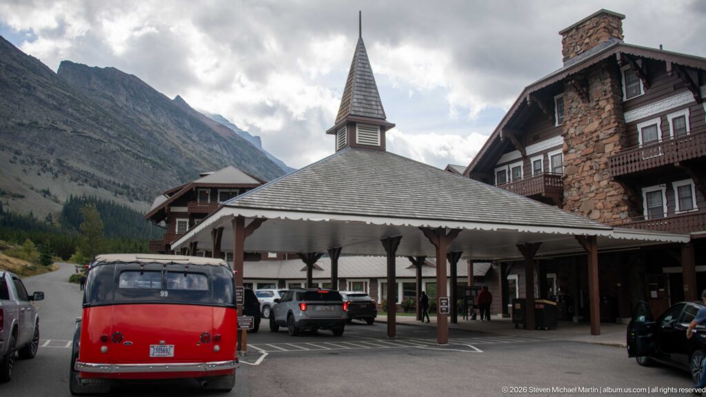 Red touring bus facing the covered Many Glacier Hotel entrance, with cars, stone chimney, balconies, and mountains nearby.