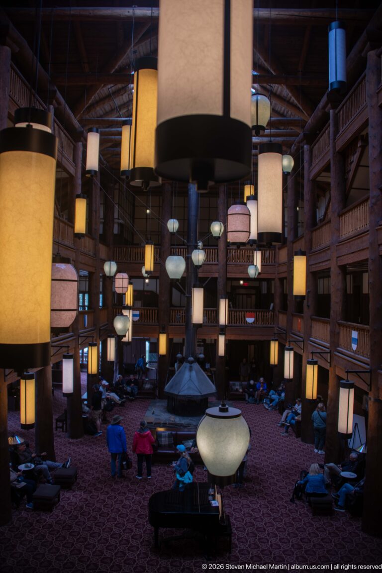 Large hanging lanterns above the Great Room, with log balconies, central fireplace, and visitors below.