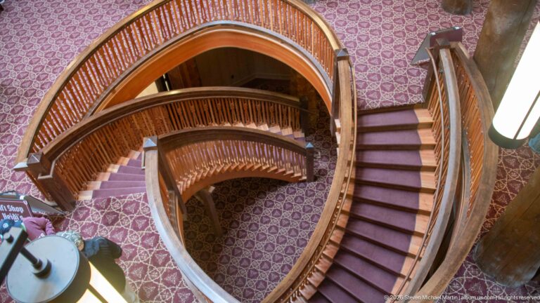 Curving wooden staircase seen from above, with rounded landings, close-set balusters, and patterned carpet.