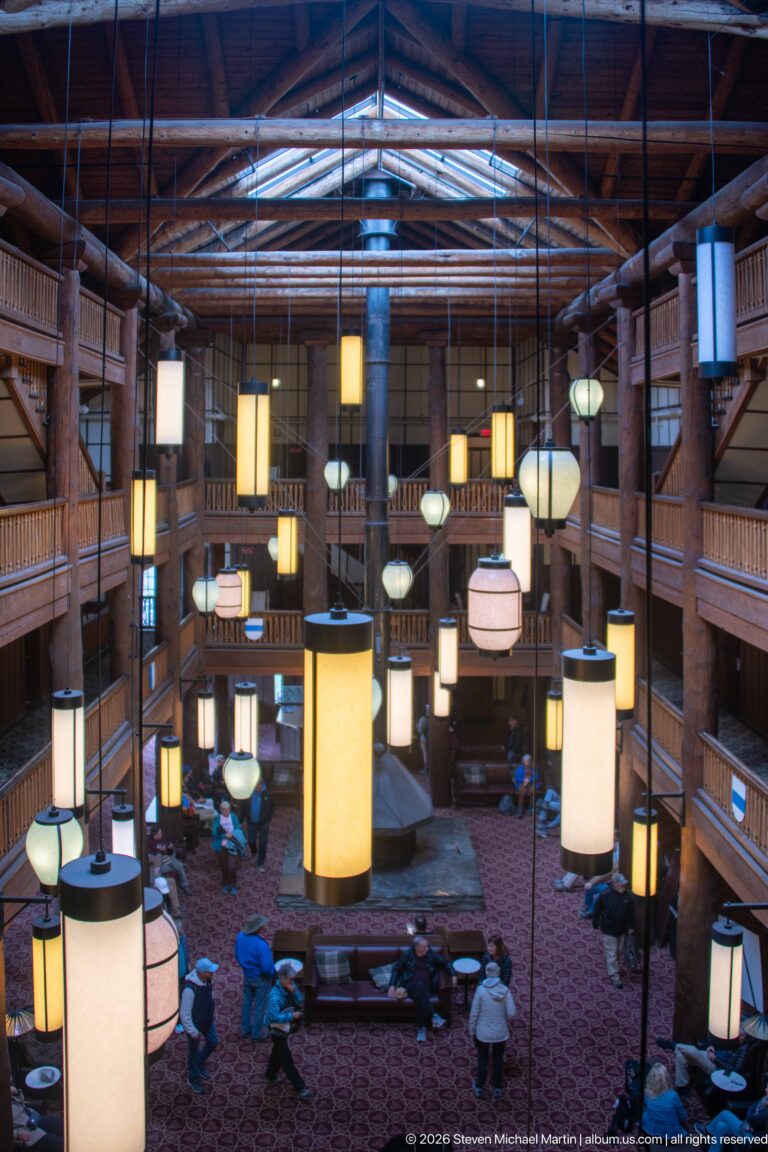 High view over the Great Room with hanging lanterns, multi-level log balconies, central fireplace hood, and seated guests.