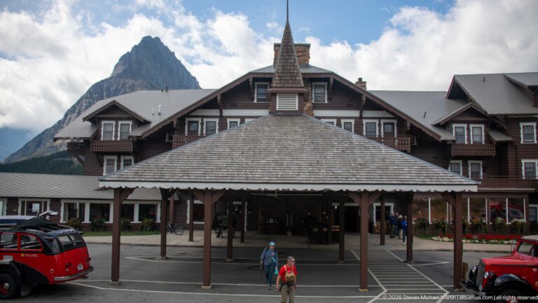 Covered entrance of Many Glacier Hotel with lodge wings behind, red touring buses, visitors, and a steep mountain peak.