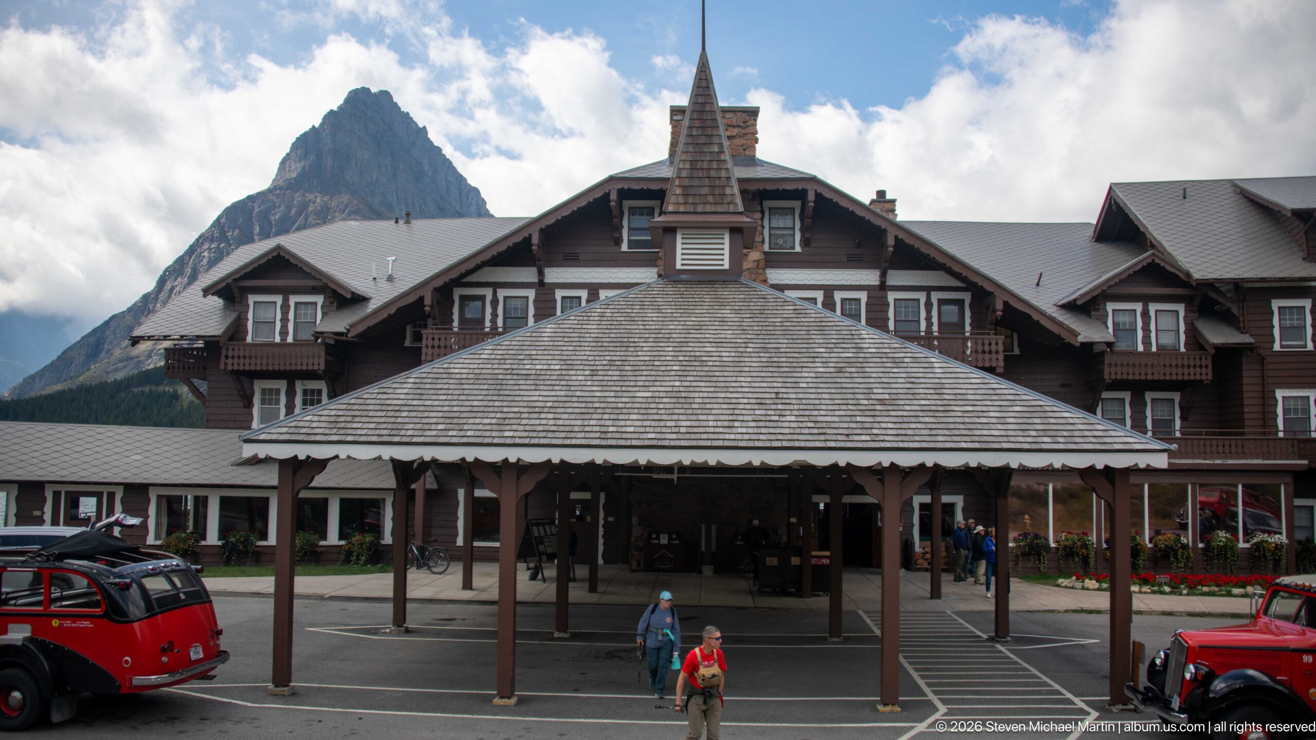Covered entrance of Many Glacier Hotel with lodge wings behind, red touring buses, visitors, and a steep mountain peak.