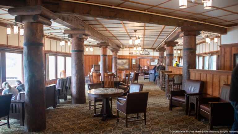 Leather chairs and small tables arranged between heavy log columns beside tall windows in the Interlaken Lounge.