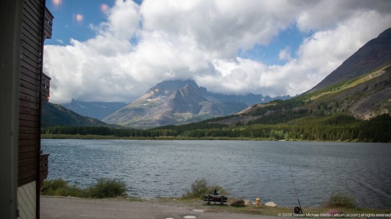 Swiftcurrent Lake and forested mountain slopes beneath broken clouds, viewed from the edge of Many Glacier Hotel.