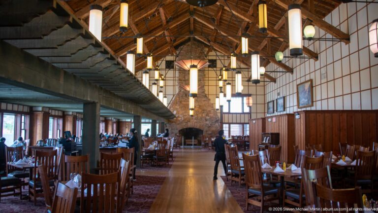 Staff member walking through the dining room between rows of wooden tables beneath hanging lanterns.