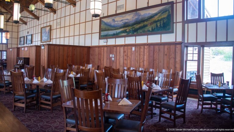Dining tables with folded napkins beside wood-paneled walls, mountain artwork, and tall windows.
