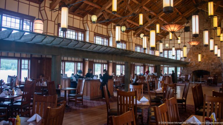 Wooden tables and chairs beneath hanging lights in the Ptarmigan Dining Room, with a stone fireplace at the far end.