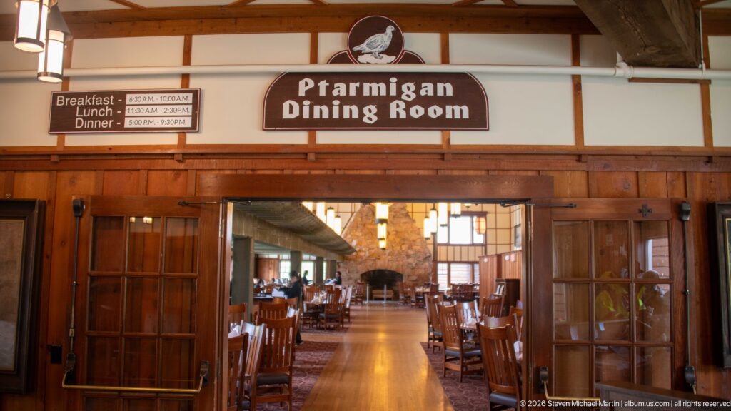 Entrance to the Ptarmigan Dining Room with wooden doors, dining tables, hanging lights, and a stone fireplace beyond.