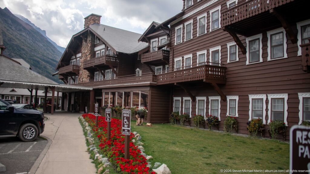 Brown Many Glacier Hotel façade with projecting wooden balconies, white-trimmed windows, stone chimney, lawn, and red flowers.