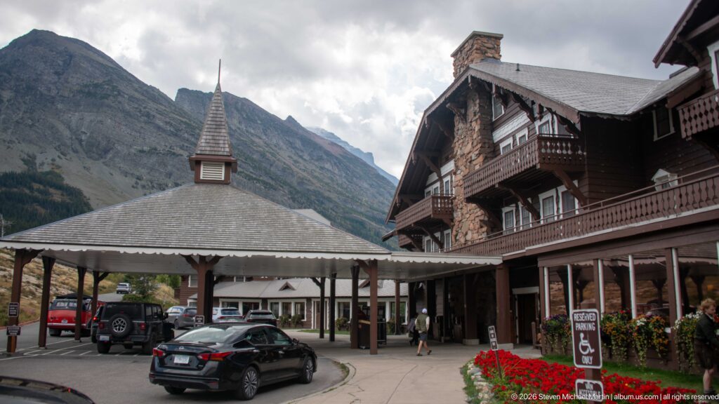 Many Glacier Hotel entrance court with covered porte-cochère, brown lodge façade, parked cars, and mountains behind.