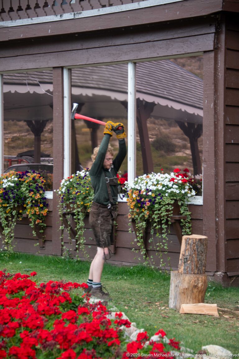 Staff member raising a red-handled splitting tool over a chopping block beside colorful lodge flower beds.