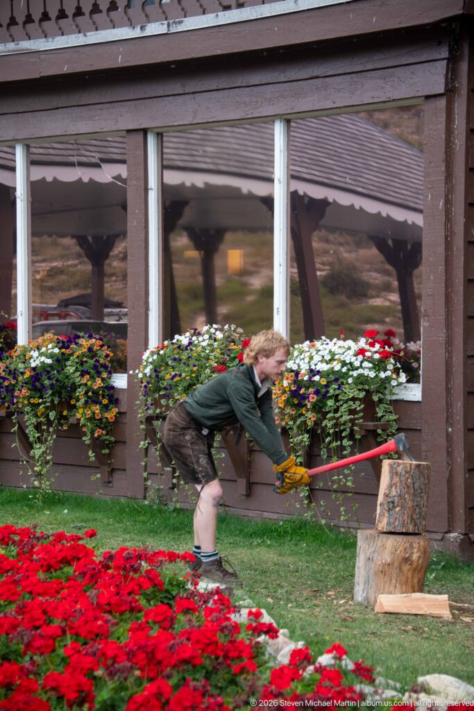 Staff member bending over a chopping block while splitting wood beside red flowers and window boxes.