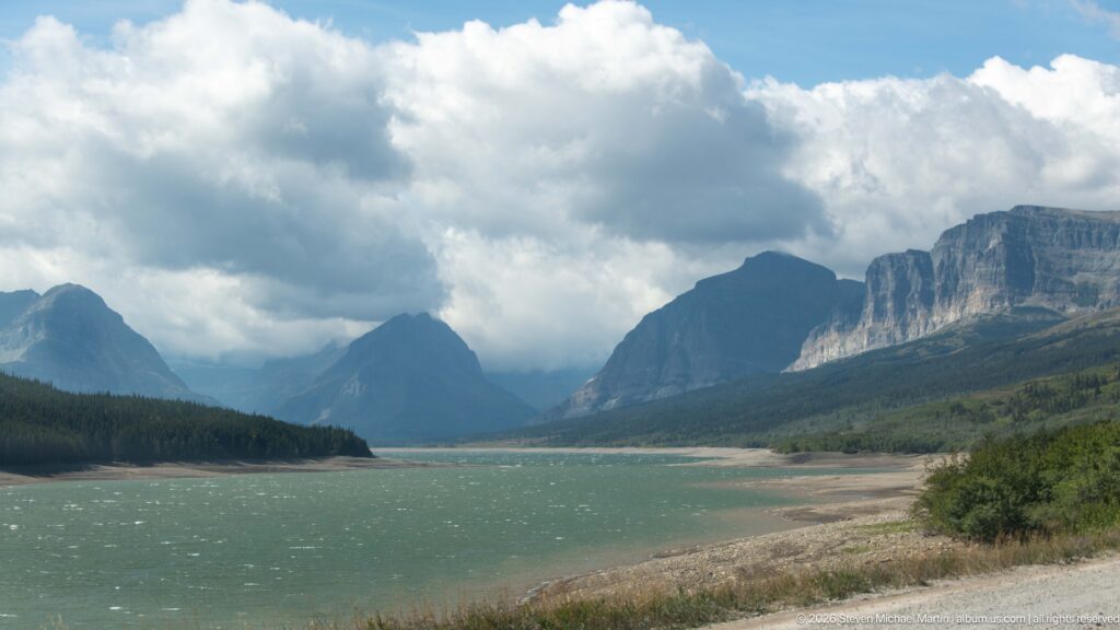Turquoise Sherburne Lake bordered by exposed shoreline and forest, with steep mountains and clouds beyond.