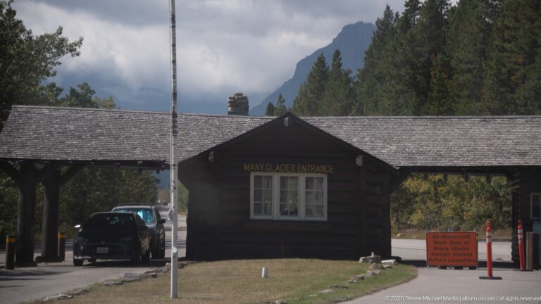 Many Glacier Entrance station spanning vehicle lanes, with dark log walls, shingled roof, car, and roadside equipment.