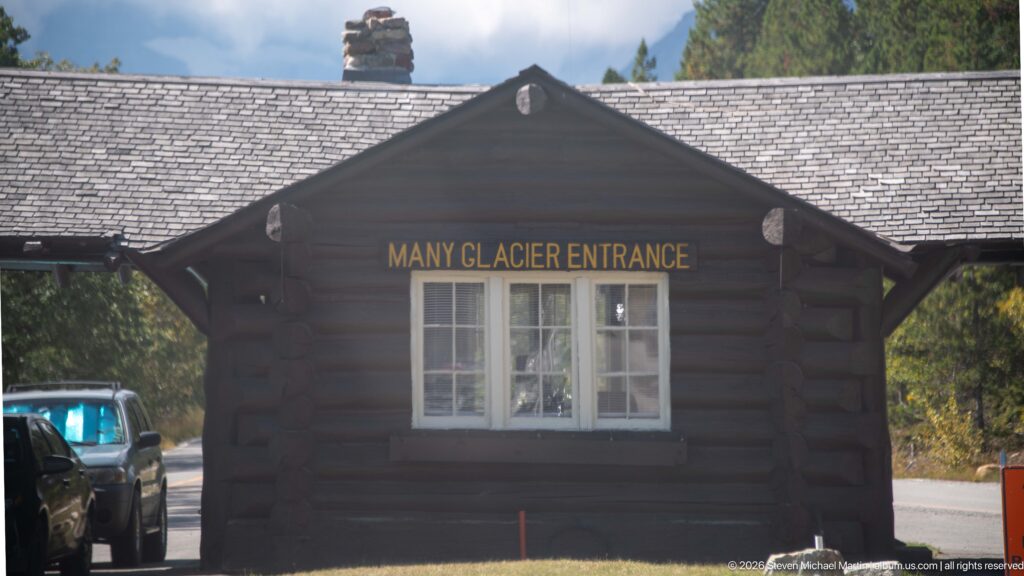 Dark log building labeled Many Glacier Entrance, with multipane windows, shingled roof, and vehicles beside it.