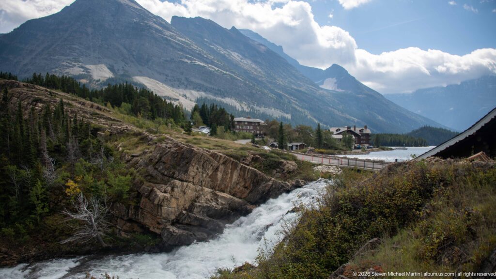 Wide valley view with a rocky cascade, roadside buildings, and a long gray mountain wall.