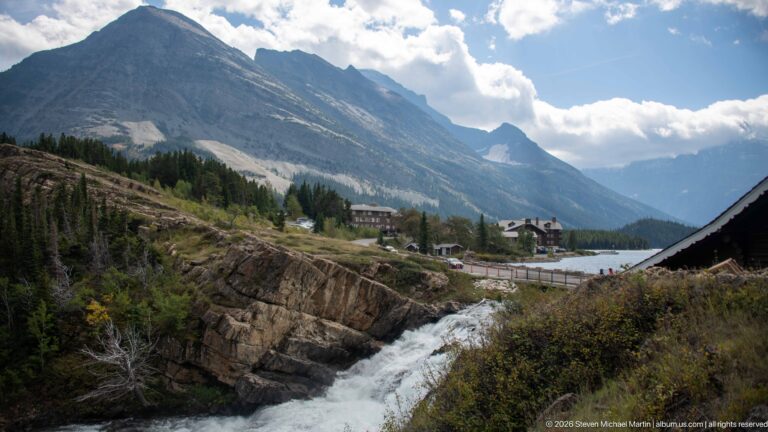 Whitewater cascade below roadside buildings and a long steep mountain wall under clouds.