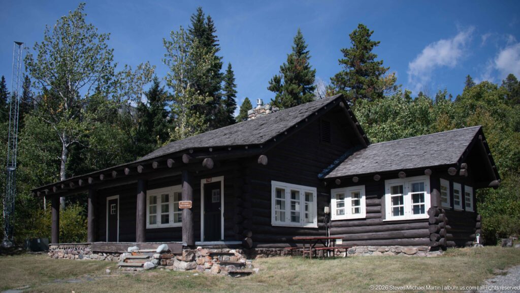 Oblique view of dark log building with intersecting shingled roofs, porch, stone base, and white windows.