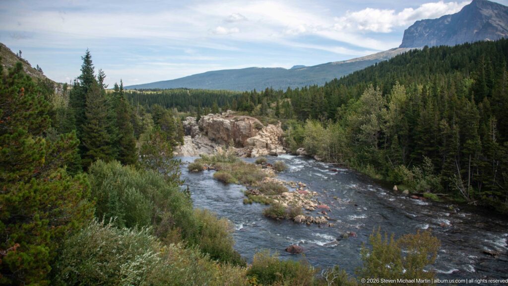 Broad rocky river flowing through conifer forest beneath layered mountain ridges.