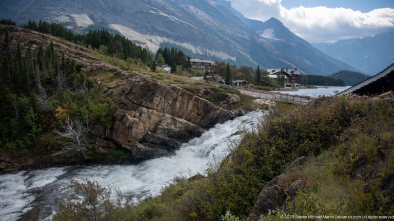 Rocky cascade below a roadside cluster of buildings with steep gray mountains behind.