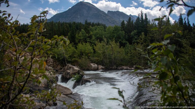 River bend and rocky bank seen through leafy branches, with forest and a steep mountain beyond.