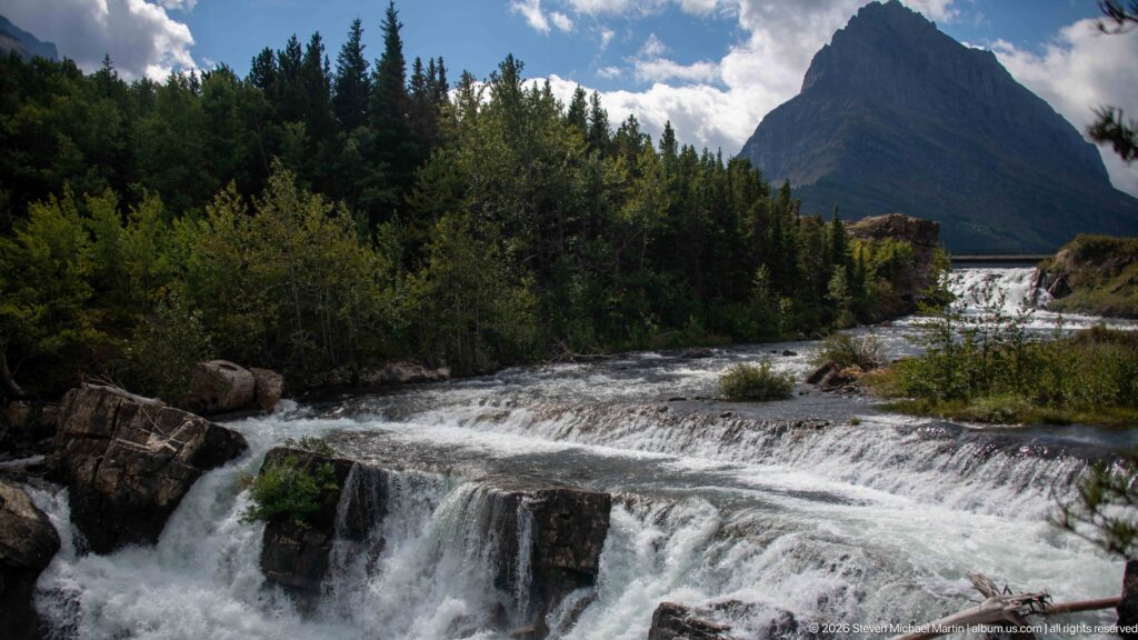 Stepped whitewater cascade below dense forest and a steep pointed mountain.