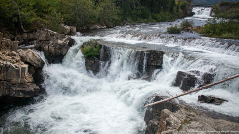Whitewater cascade dropping over dark rock ledges with a fallen trunk across the lower flow.