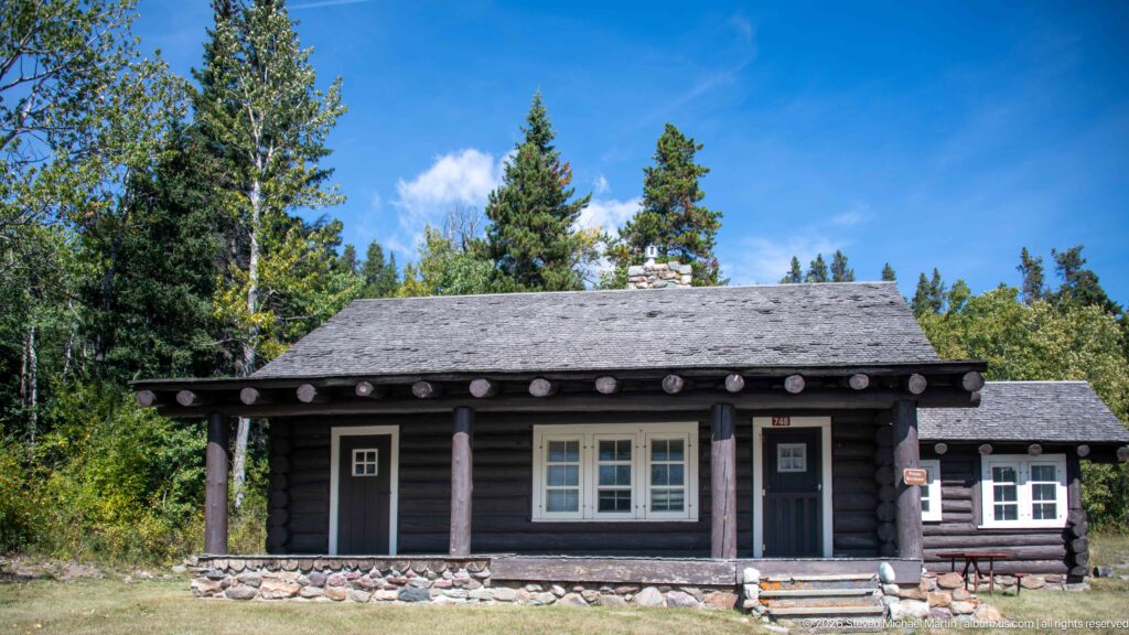 Dark log entrance-area building with shingled roof, log porch posts, stone base, and white-trimmed windows.