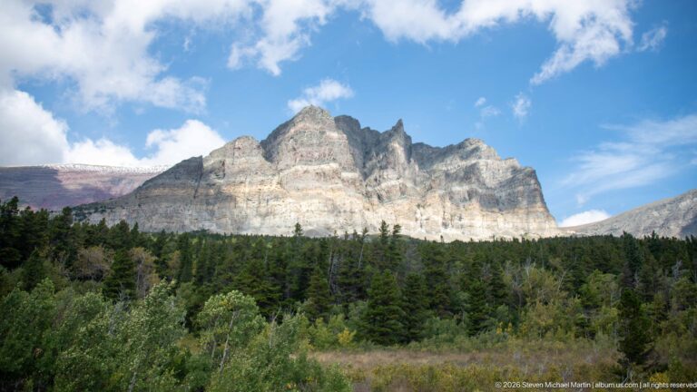 Pale layered mountain ridge with a jagged crest rising above dark conifer forest under blue sky and clouds.