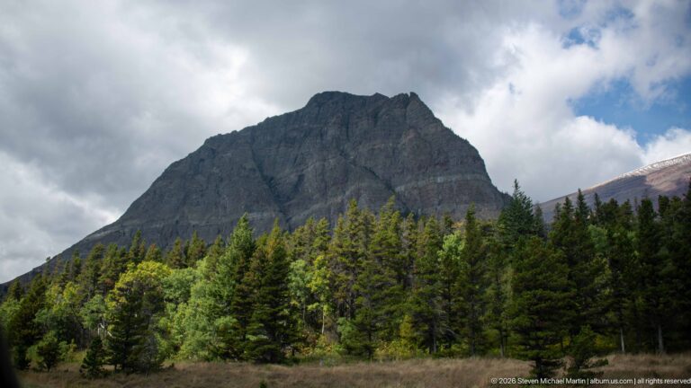 Steep dark mountain rising above mixed forest beneath gray and white clouds along Many Glacier Road.