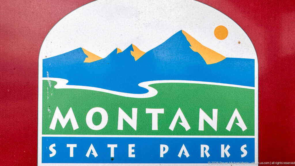 Montana State Parks logo with blue mountains, green foreground, white winding line, and bold lettering on red.