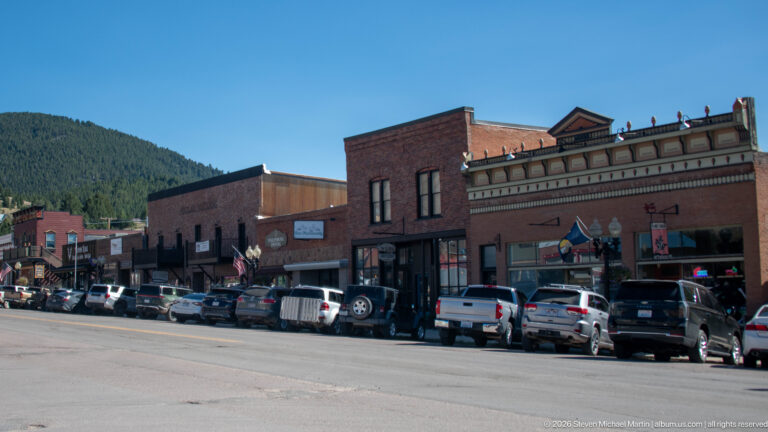 Row of historic Philipsburg storefronts and parked vehicles along a broad street with a forested mountain rising behind downtown.