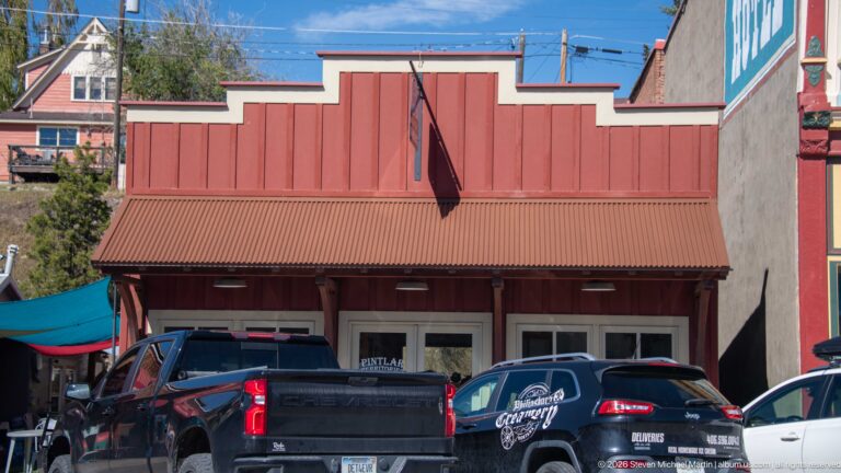 Red stepped false-front storefront with vertical siding, corrugated metal awning, flag, display windows, and parked vehicles.