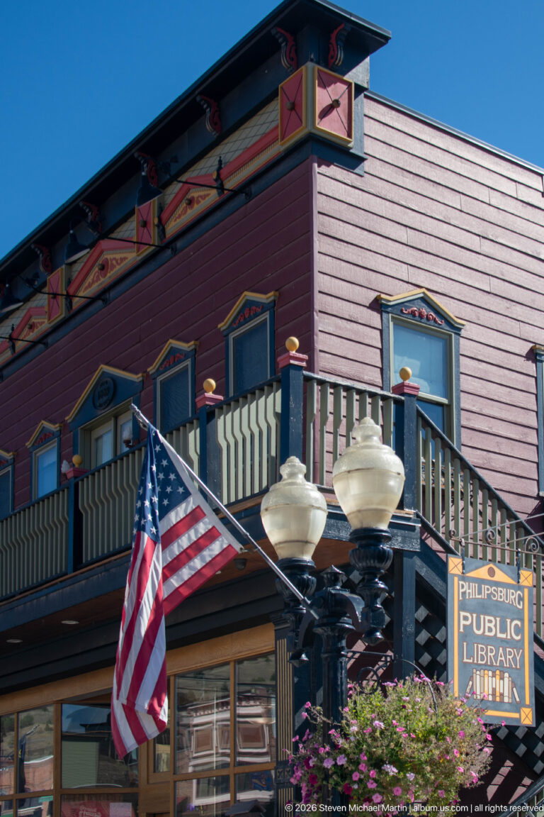 Philipsburg Public Library in a two-story painted wood building with wraparound balcony, decorative trim, American flag, and streetlamps.