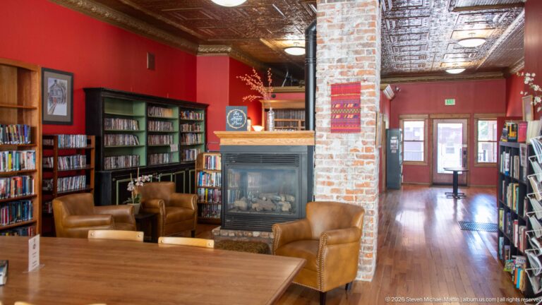Library reading room with leather chairs around a fireplace, bookcases, exposed brick pier, wood floor, and pressed-metal ceiling.