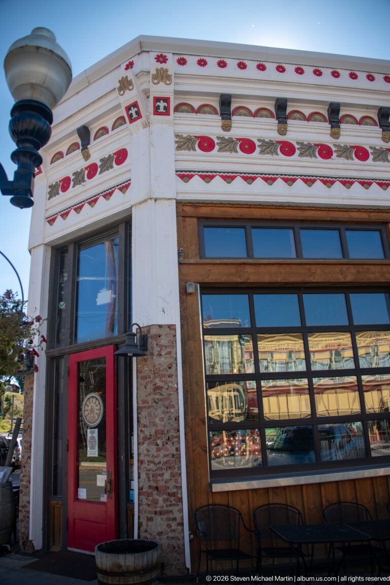 Ornate white corner storefront with red motifs, dark brackets, red door, tall windows, exposed brick, and reflected historic buildings.