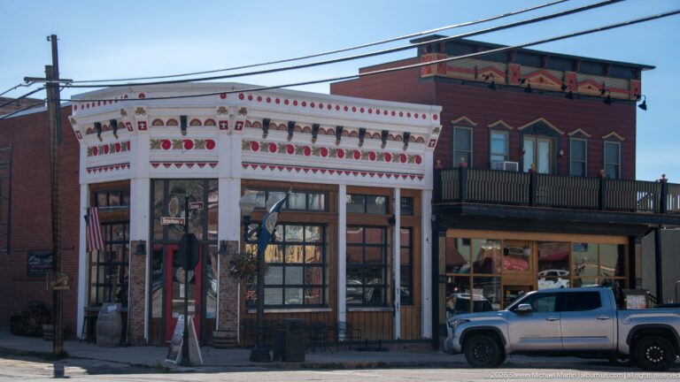 Ornate white historic storefront beside a painted two-story wood building with balcony, decorative trim, and a parked pickup.