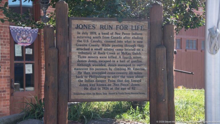 Wood-framed historical marker titled JONES’ RUN FOR LIFE beside a brick building and shaded grass in Philipsburg.