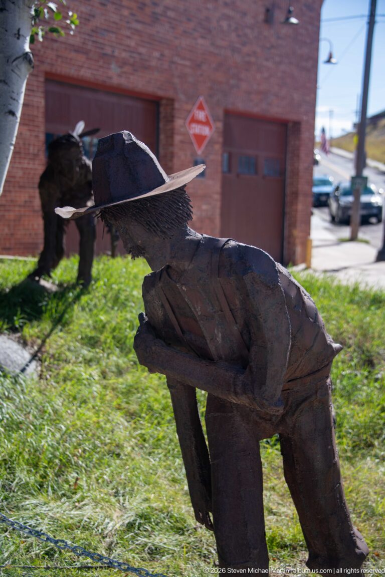 Life-size weathered metal figure bent forward on a grassy lawn, with another rustic sculpture and brick building behind.