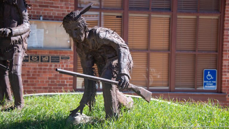 Weathered life-size metal figure leaning forward with a long rifle-like object on grass beside a brick building.