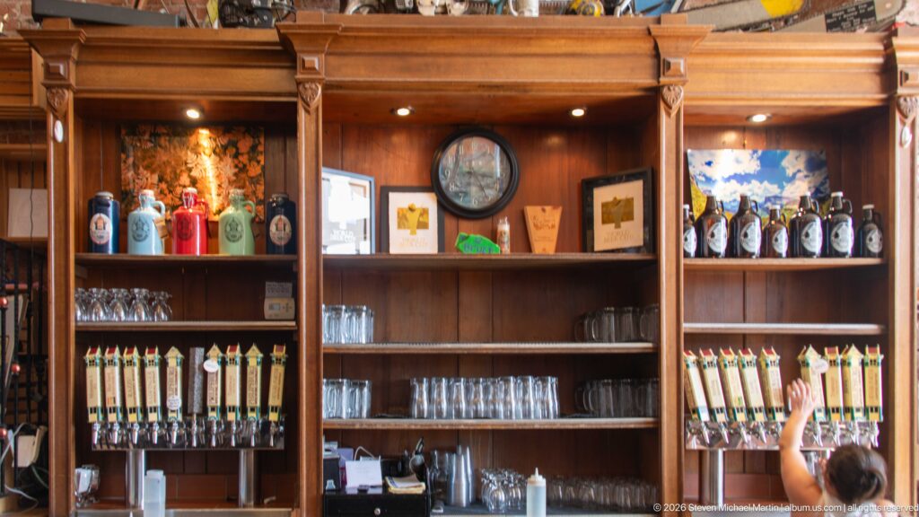 Wood bar at Philipsburg Brewing Company with rows of beer taps, glassware, bottles, and a staff member reaching toward a tap.