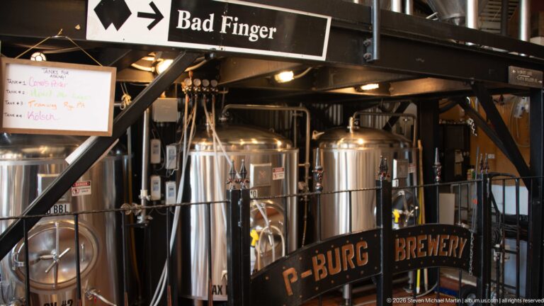 Stainless brewery tanks beneath P-BURG BREWERY and Bad Finger signs, with black steel framing, hoses, valves, and gauges.