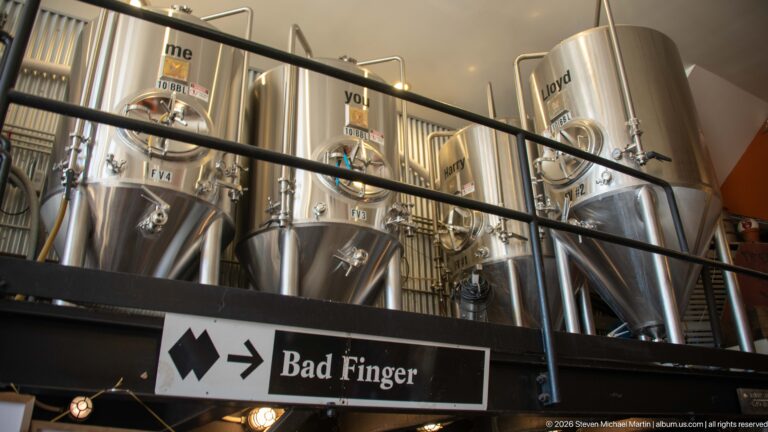 Elevated stainless brewing tanks with conical bottoms, valves, railing, and a Bad Finger directional sign below.