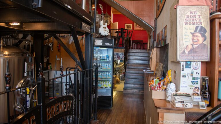 Philipsburg Brewing Company passage with stainless tanks, P-BURG BREWERY railing, cooler, merchandise, staircase, guitar, and wall poster.