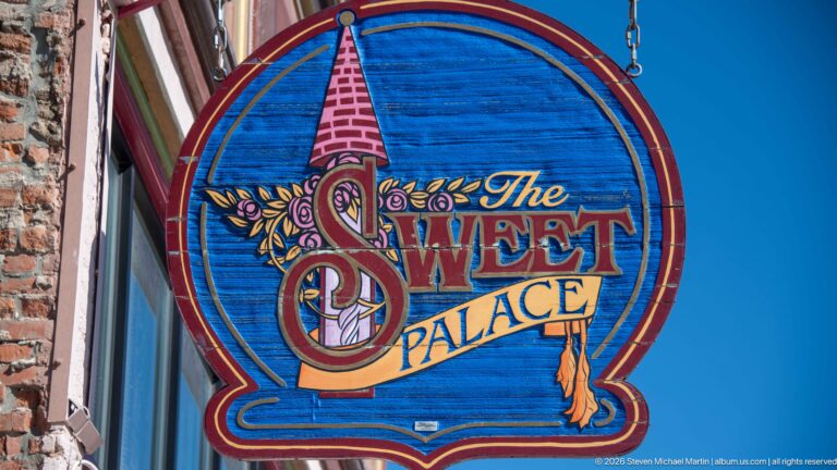 Colorful blue hanging sign for The Sweet Palace with ornate lettering, floral decoration, and a small orange cat motif.