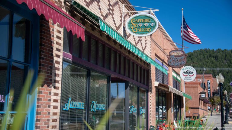 Sidewalk view past Sapphire Gallery and neighboring brick storefronts, projecting signs, American flag, and wooded mountain slope.