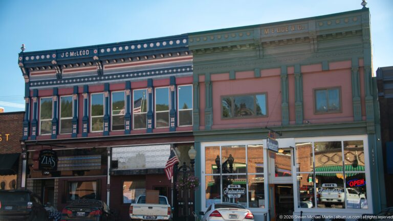 J.C. McLeod Building beside the M.E. Doe Building, showing contrasting painted façades, upper windows, storefronts, and parked vehicles.