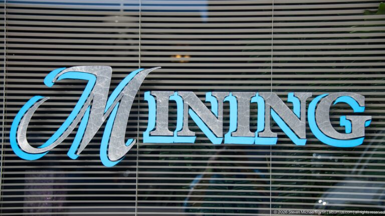Decorative blue-and-silver MINING lettering applied to a storefront window with horizontal blinds behind it.